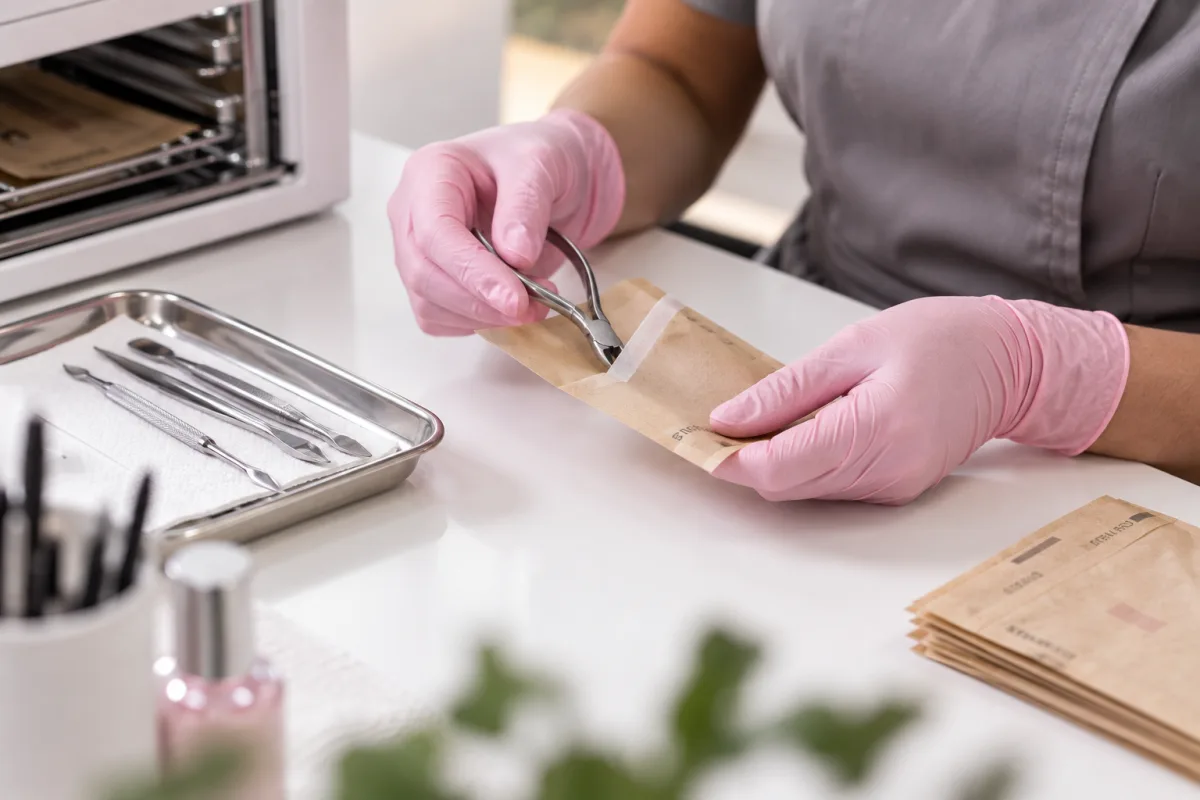 Een nagelstyliste met handschoenen verpakt gereinigde instrumenten in een sterilisatiezakje naast de autoclaaf